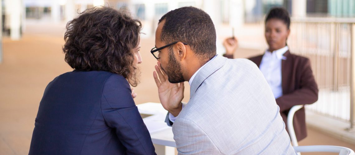Employees gossiping about young female colleague. Business man and woman whispering, African American employee sitting in background. Office rumors concept