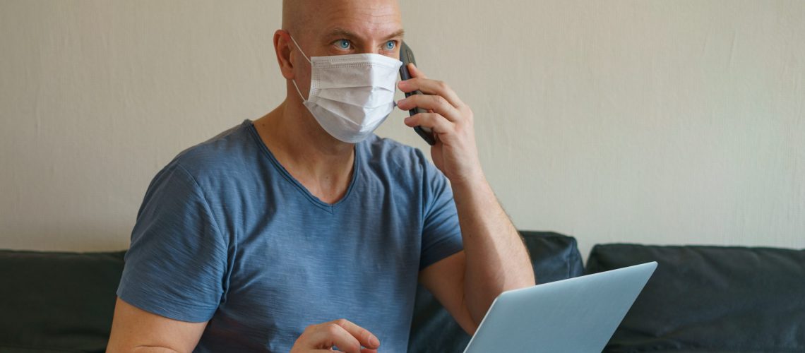 Man is sitting on a sofa in a protective mask with a laptop and phone, remote work in quarantine